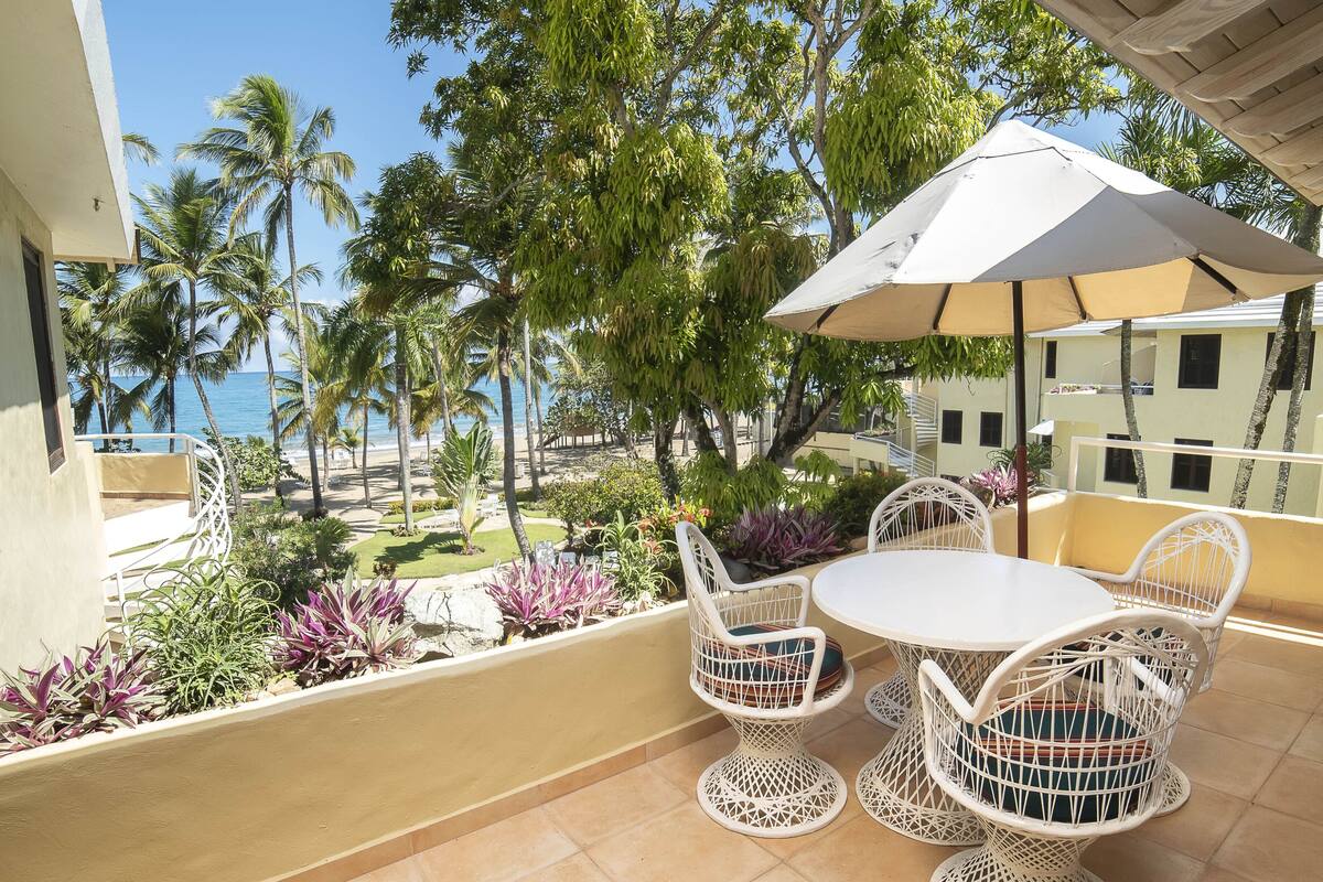 A balcony with a white table and chairs, an umbrella, and a view of the ocean and palm trees.
