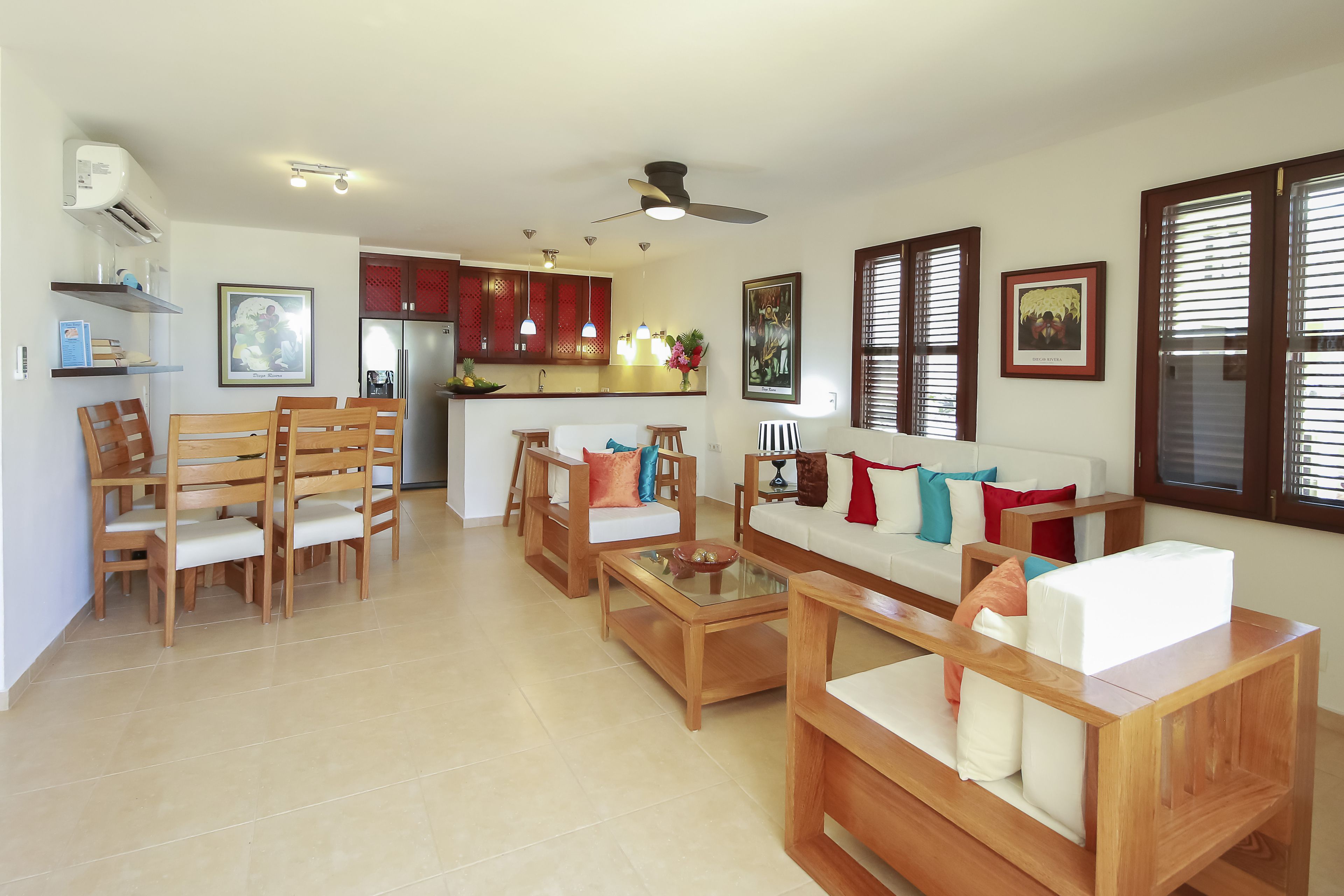 A spacious living area with a sofa, chairs, and a coffee table. There is a kitchen in the background with cabinets and a refrigerator.