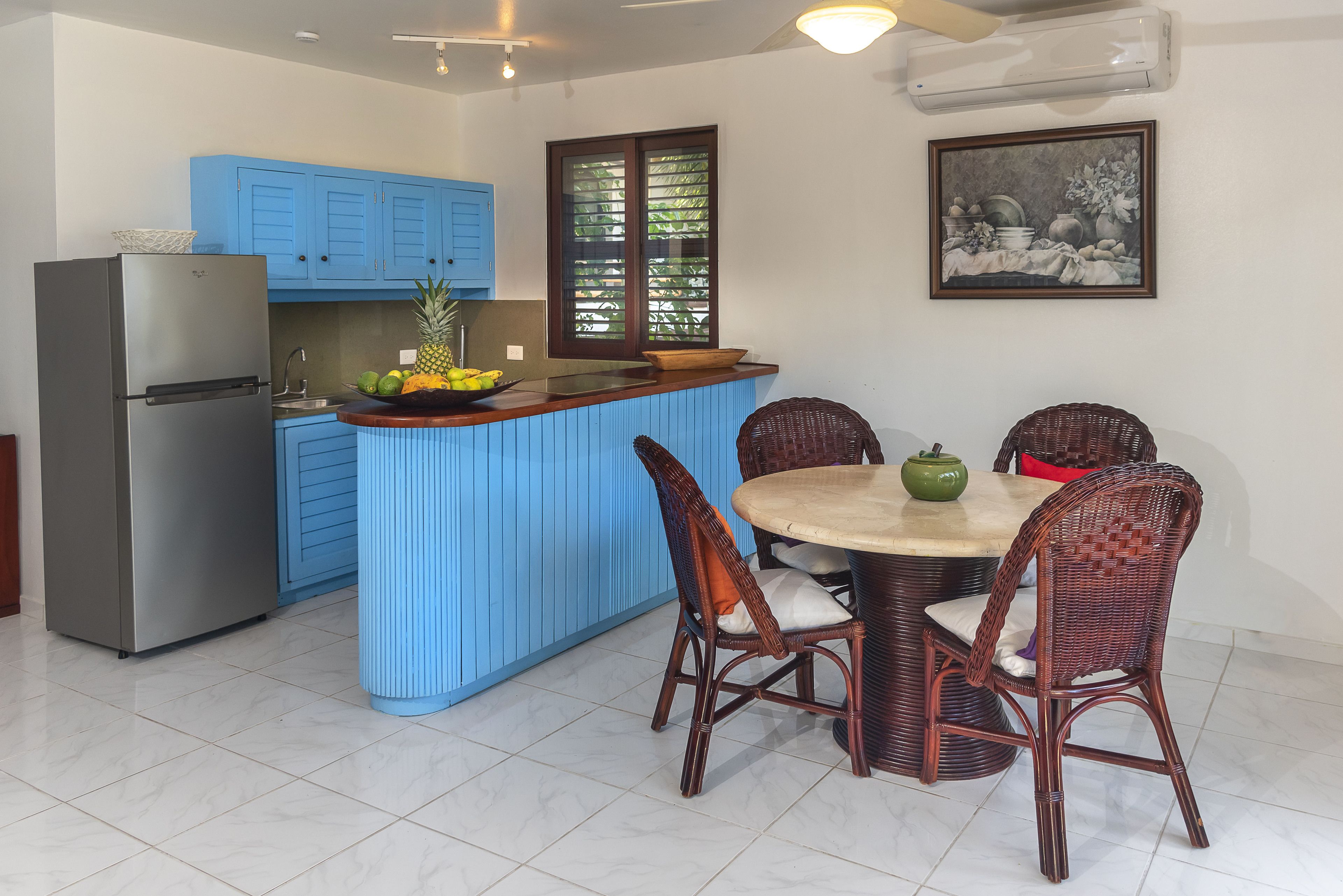 Une cuisine avec des meubles de rangement bleus, un réfrigérateur, une table à manger avec des chaises en osier et une fenêtre donnant sur un paysage verdoyant.