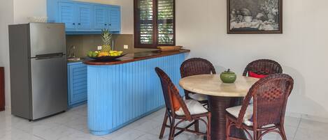 Une cuisine avec des meubles de rangement bleus, un réfrigérateur, une table à manger avec des chaises en osier et une fenêtre donnant sur un paysage verdoyant.