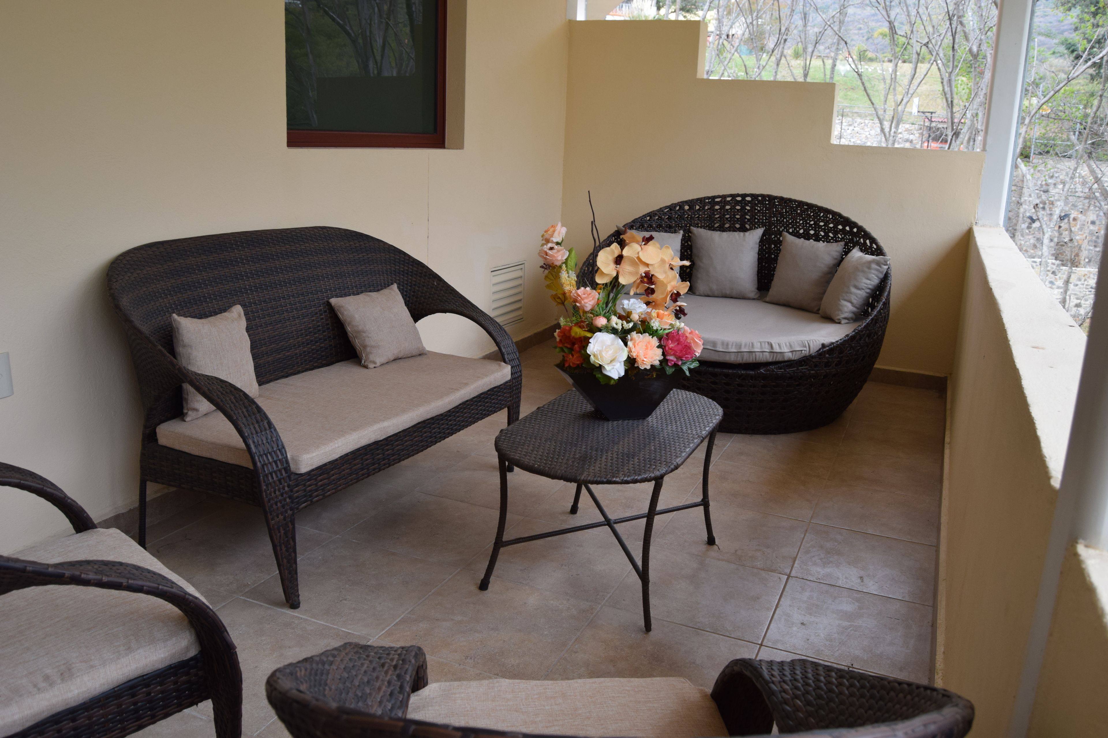 Un balcón con muebles de mimbre, incluyendo un sofá y sillas, y una pequeña mesa con un arreglo floral.