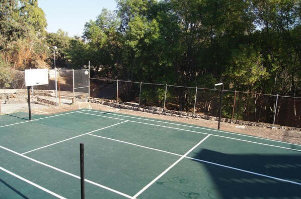 Una cancha de tenis al aire libre con red, rodeada por una valla de malla metálica y árboles.