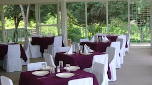 A covered outdoor dining area with tables set for a formal event, surrounded by white chairs and draped with maroon tablecloths.