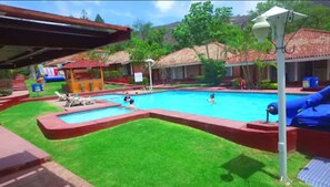 Una piscina con tobogán, un área de juegos pequeña y una zona de estar cubierta.