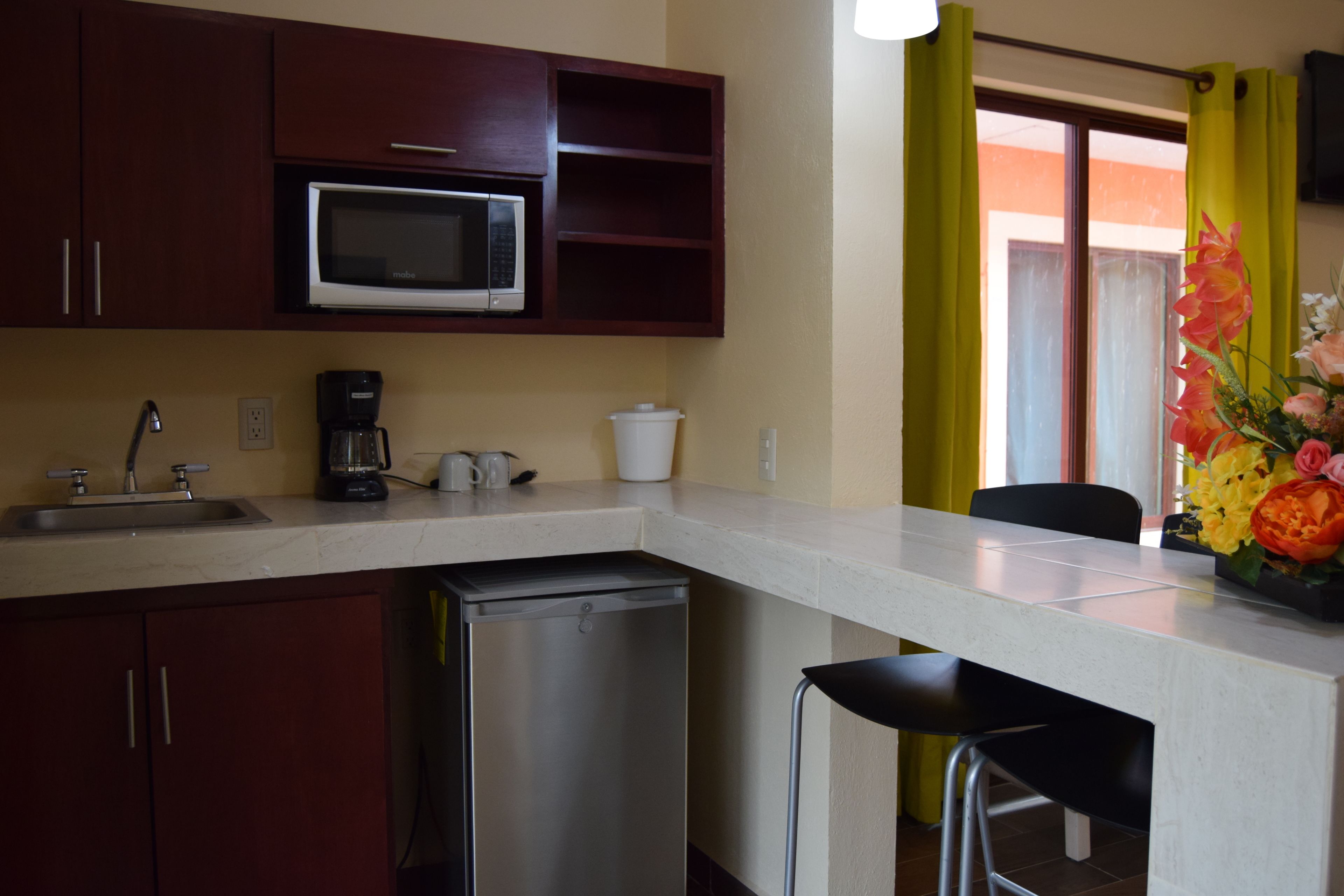 A compact kitchen with a microwave, coffee maker, and a small refrigerator. There is a sink with a faucet, and a chair is visible near the window.