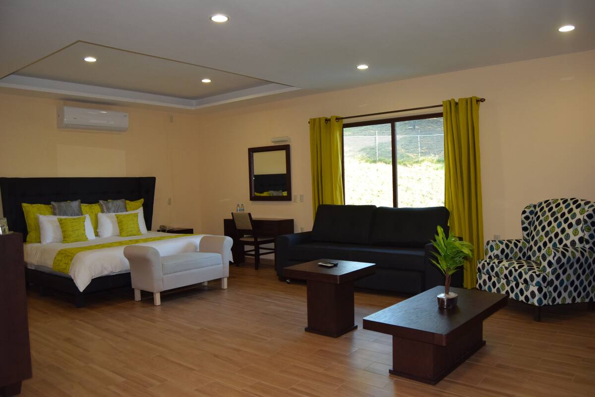 Habitación de hotel con cama, sofá, mesa de centro, sillón estampado y ventana con cortinas amarillas.