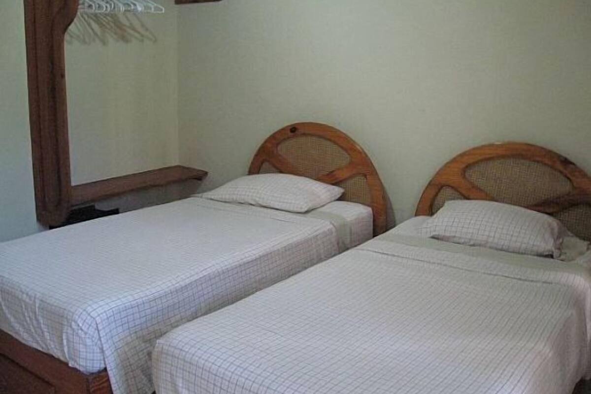 Deux lits simples avec des têtes de lit en bois, dans une pièce dont un mur est uni et qui est équipée d’une étagère en bois.