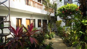 Un patio con un sendero de piedra, rodeado de plantas diversas y un edificio blanco con balcones.