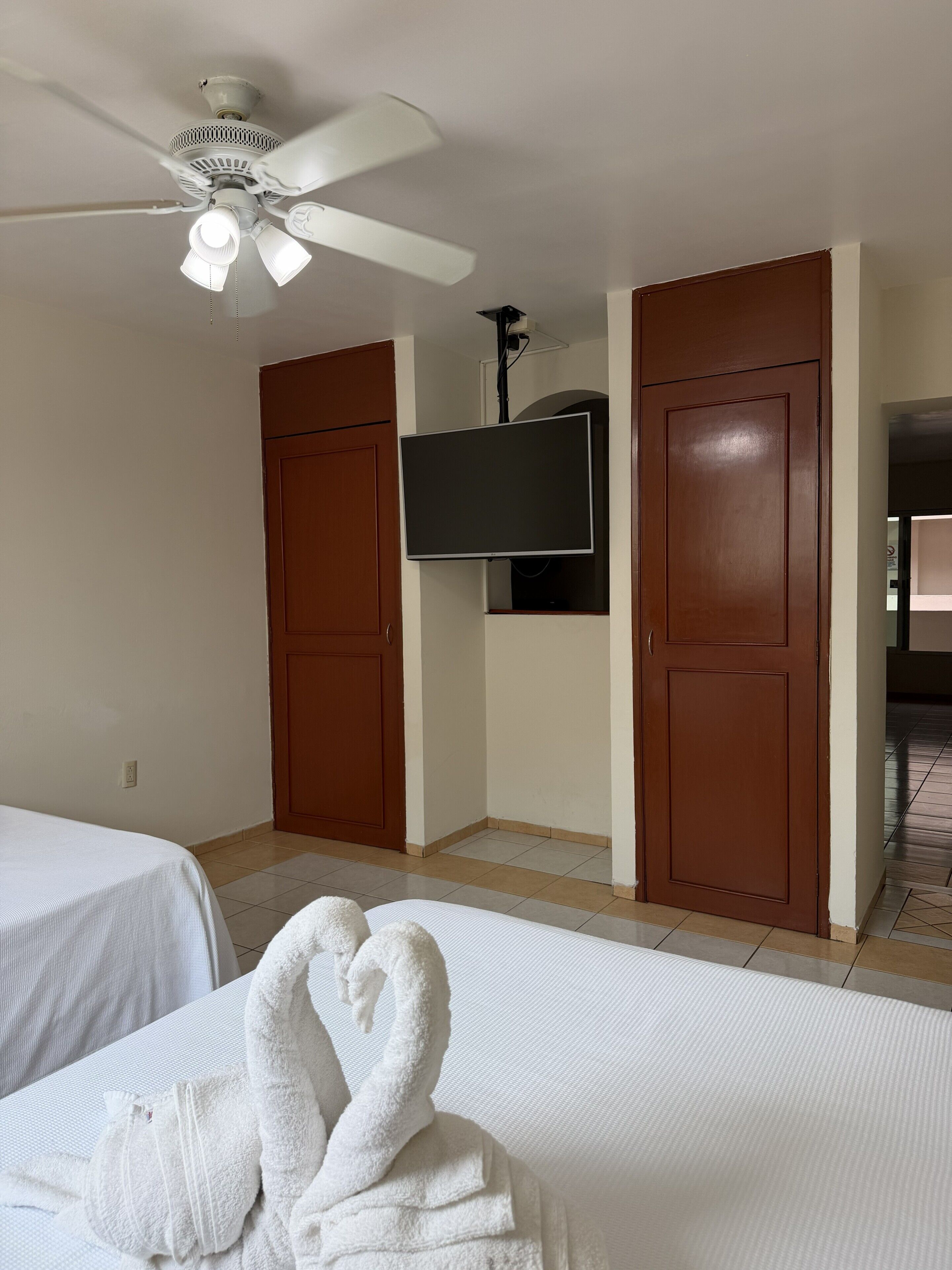 A hotel room with a bed, a ceiling fan, a TV mounted on the wall, and two wooden doors.