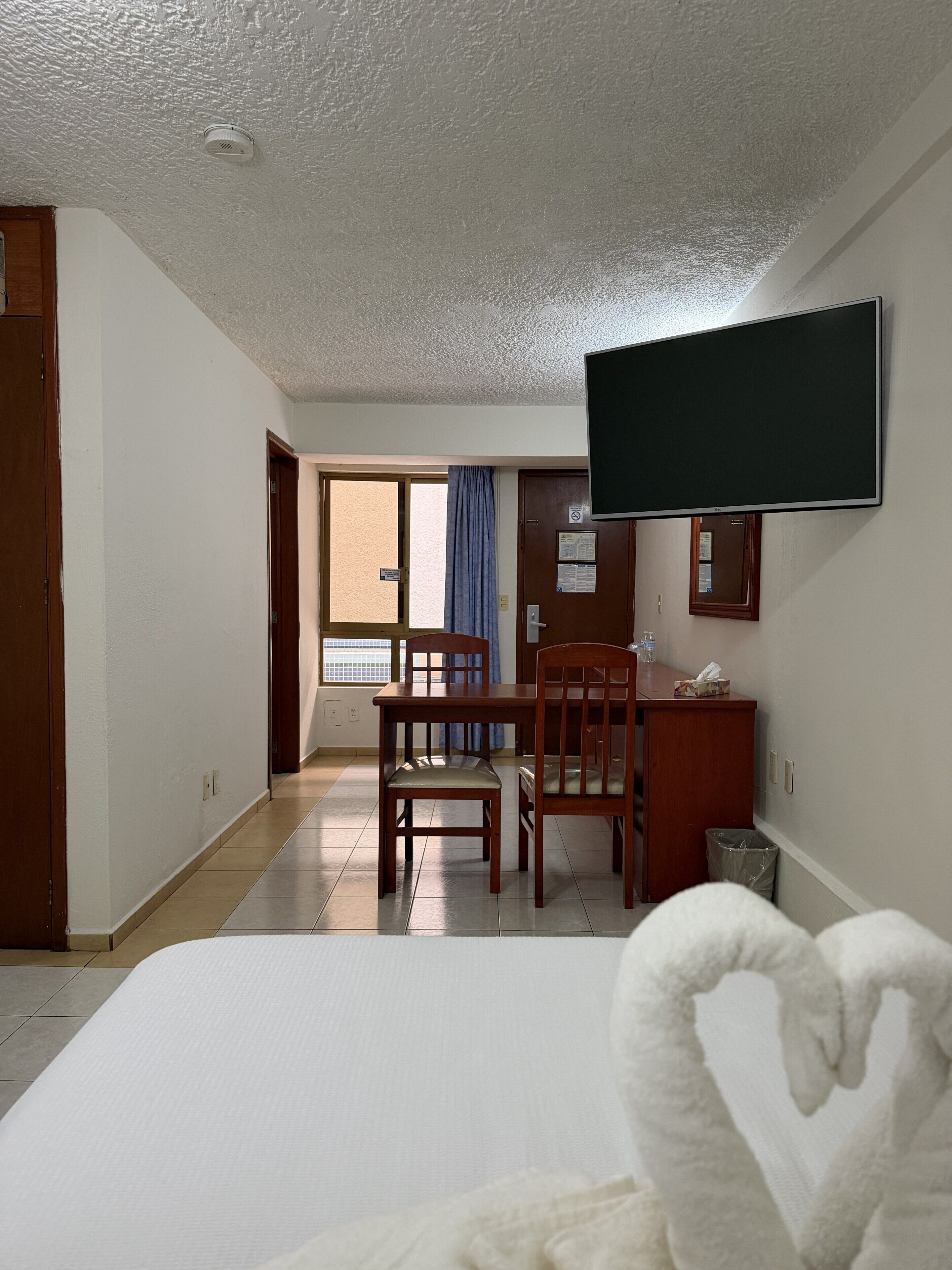 A hotel room with a bed, a desk with a chair, a television mounted on the wall, and a window with curtains.