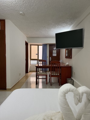 A hotel room with a bed, a desk with a chair, a television mounted on the wall, and a window with curtains.