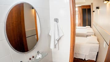 Un baño de hotel con un espejo redondo, un lavamanos y una ducha. Una puerta de madera da paso a un dormitorio con dos camas.