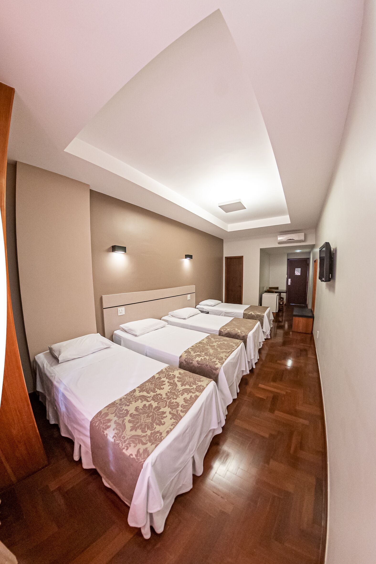 A hotel room with four beds aligned in a row, a wooden floor, and a corridor leading to a door.