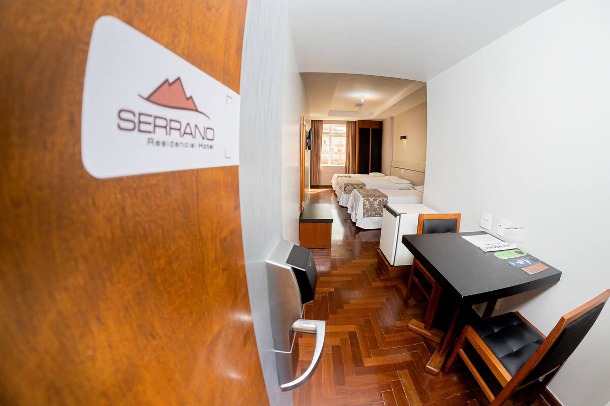 Habitación de hotel con piso de madera, una cama con cabecera blanca, una pequeña mesa con sillas y un cartel en la pared.