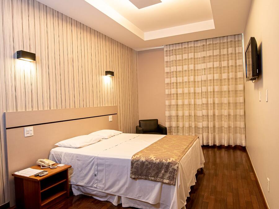 Quarto de hotel com cama, mesa de cabeceira, abajur e televisão na parede.