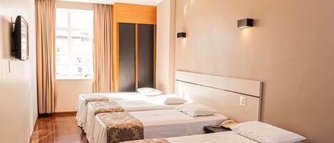 Habitación de hotel con tres camas, un ventanal con cortinas, un televisor de pantalla plana en la pared y una lámpara en el techo.