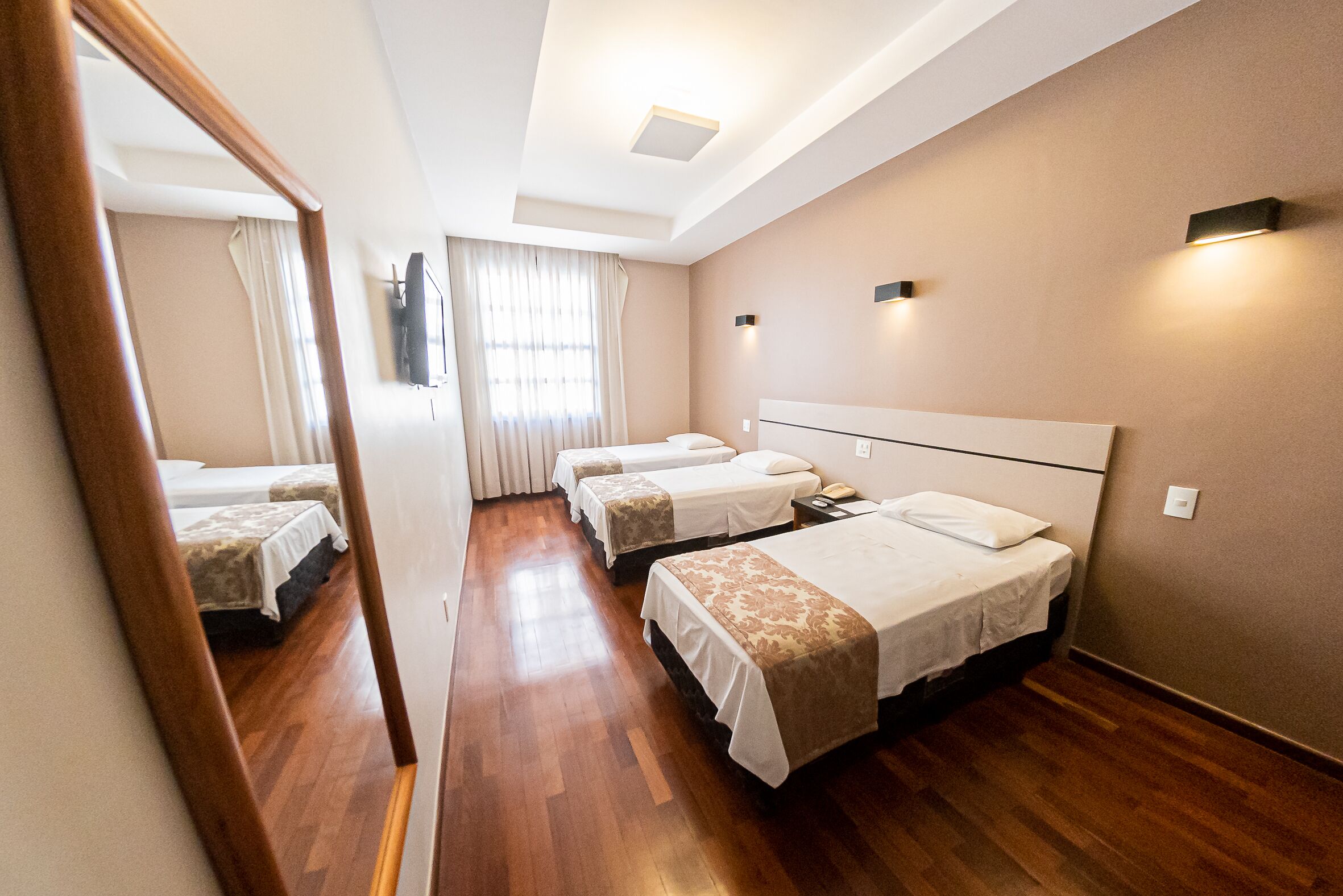 Hotellihuone, jossa on kolme erillistä sänkyä, puulattia, suuri peili ja kattovalaisin.