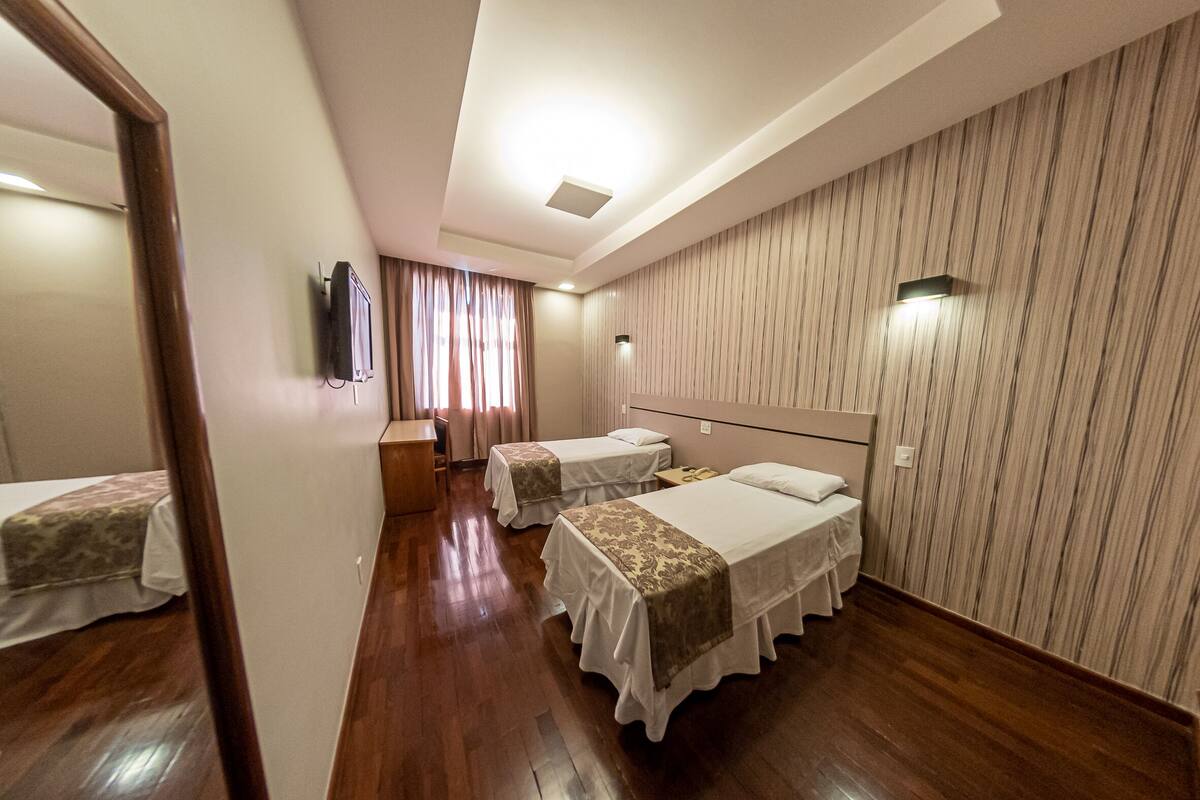 Habitación de hotel con dos camas individuales, suelo de madera, un espejo, un televisor y una ventana con cortinas.