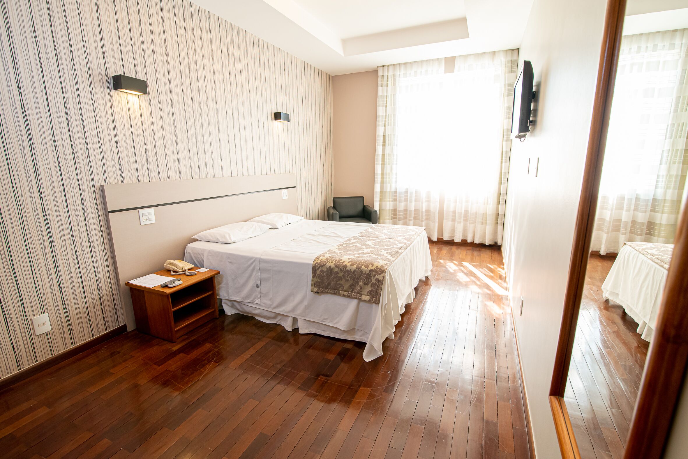 Una habitación de hotel con una cama grande, una mesita de noche con teléfono, una silla y un televisor fijado a la pared.