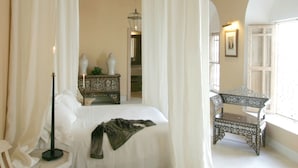 Un dormitorio con cama, cortinas blancas, lámpara colgante, mesita de noche con lámpara y ventana con vistas.