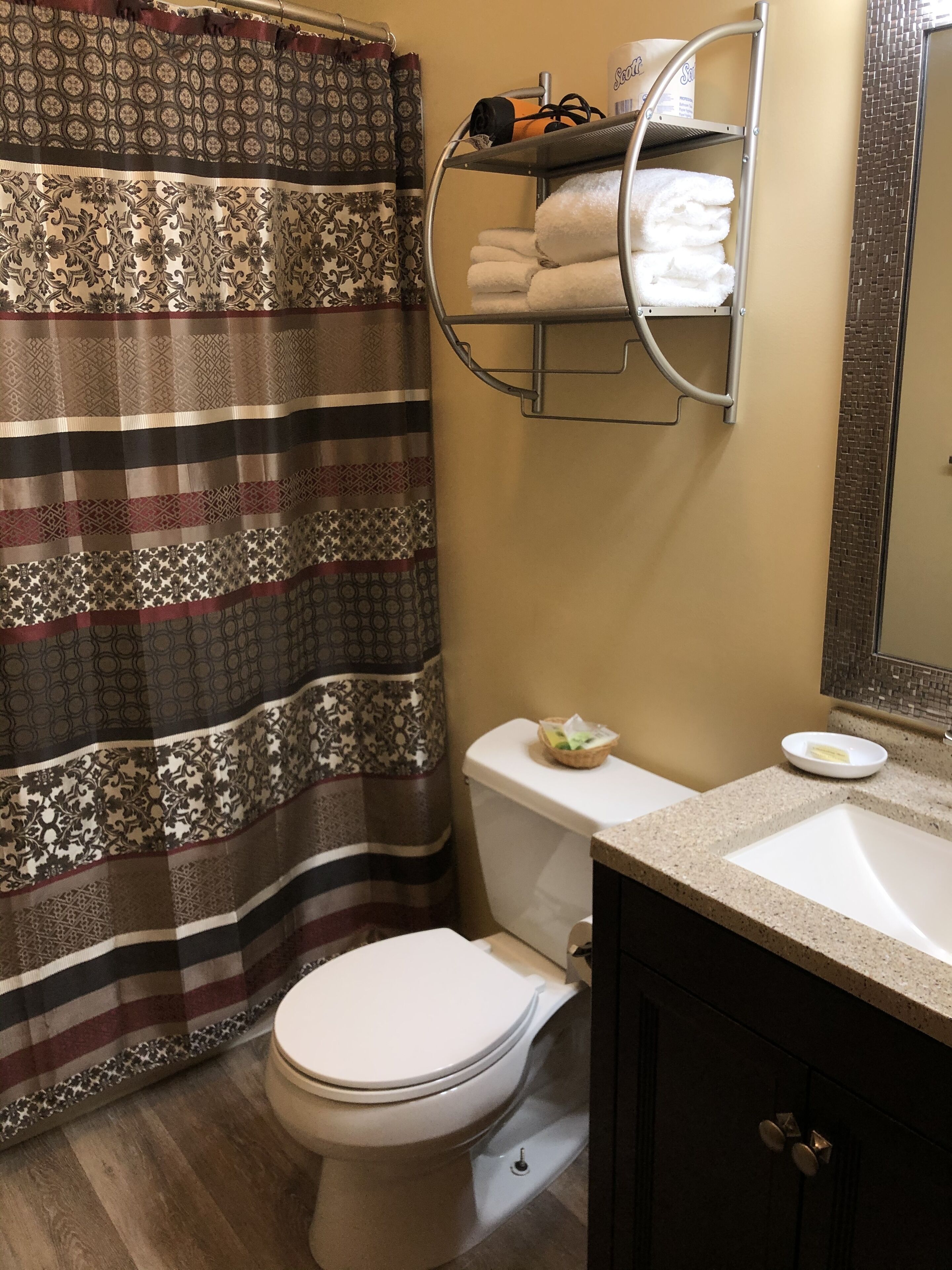 Une salle de bain avec un rideau de douche à motifs, des toilettes, un porte-serviettes avec des serviettes pliées, un porte-savon et un miroir.