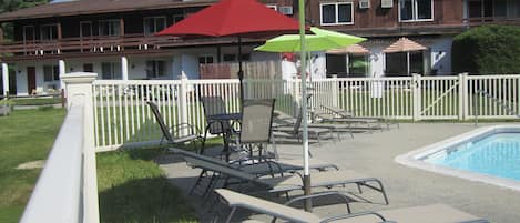 A poolside area with lounge chairs and umbrellas, a pool, and a building with a white fence in the background.