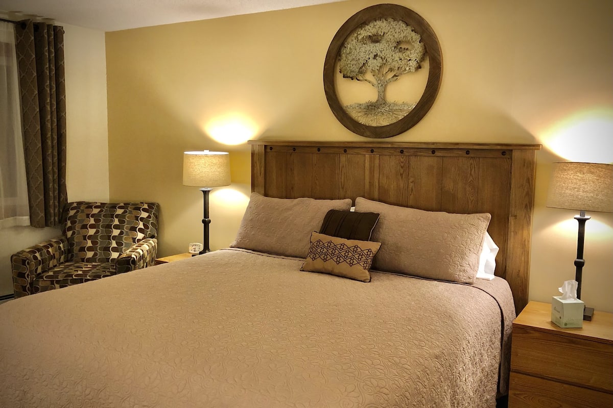 A hotel room with a large bed, two bedside tables with lamps, a patterned armchair, and a wall-mounted artwork of a tree.