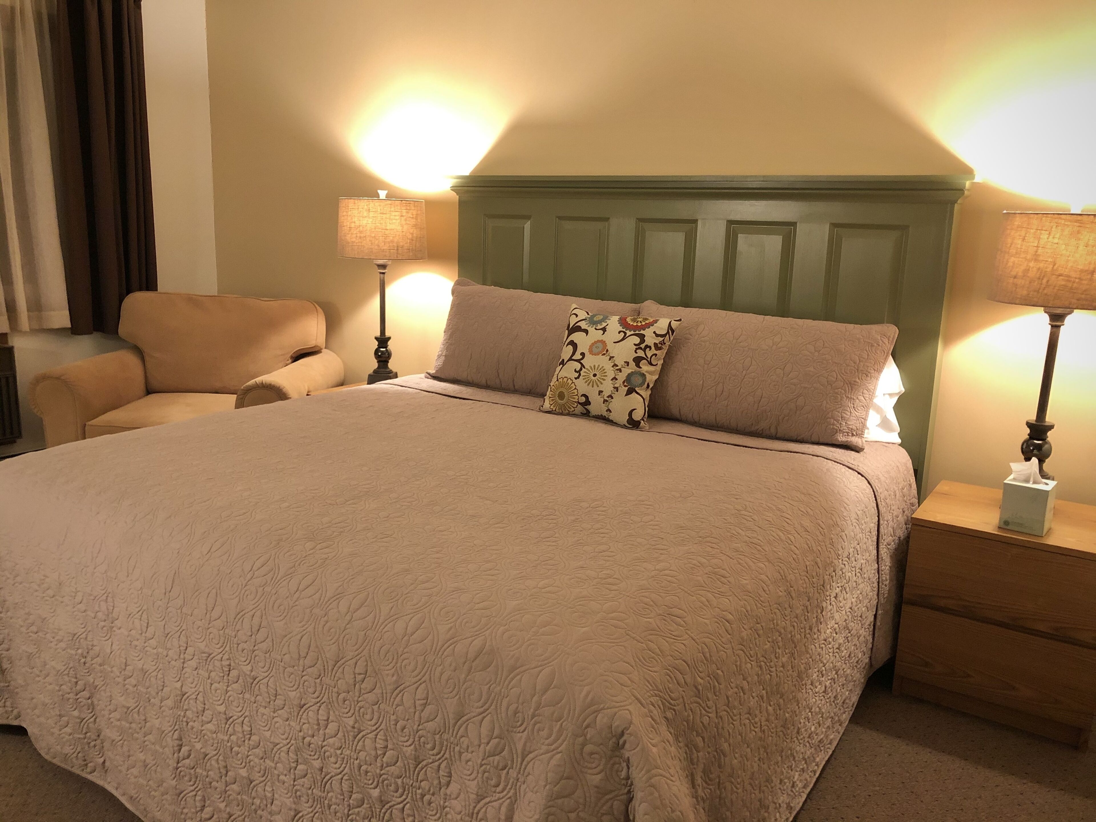 A neatly made bed with a beige quilt, two bedside lamps, a wooden nightstand, and a chair.