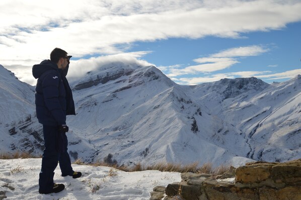 Une personne se tient debout sur une montagne enneigée, dont le terrain est rocheux, sous un ciel dégagé.