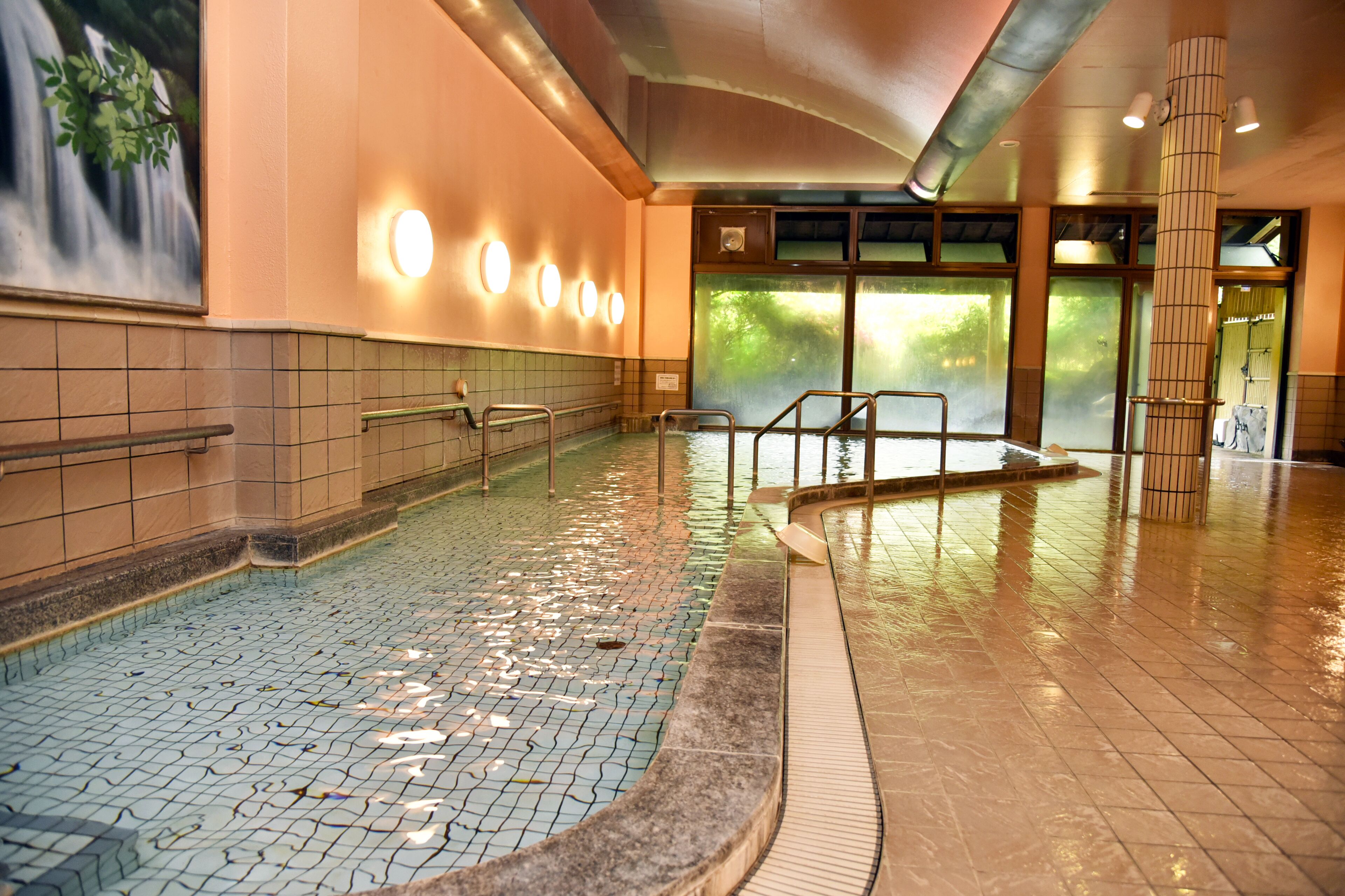 An indoor swimming pool with tiled flooring, a handrail, and a wall-mounted light fixture.