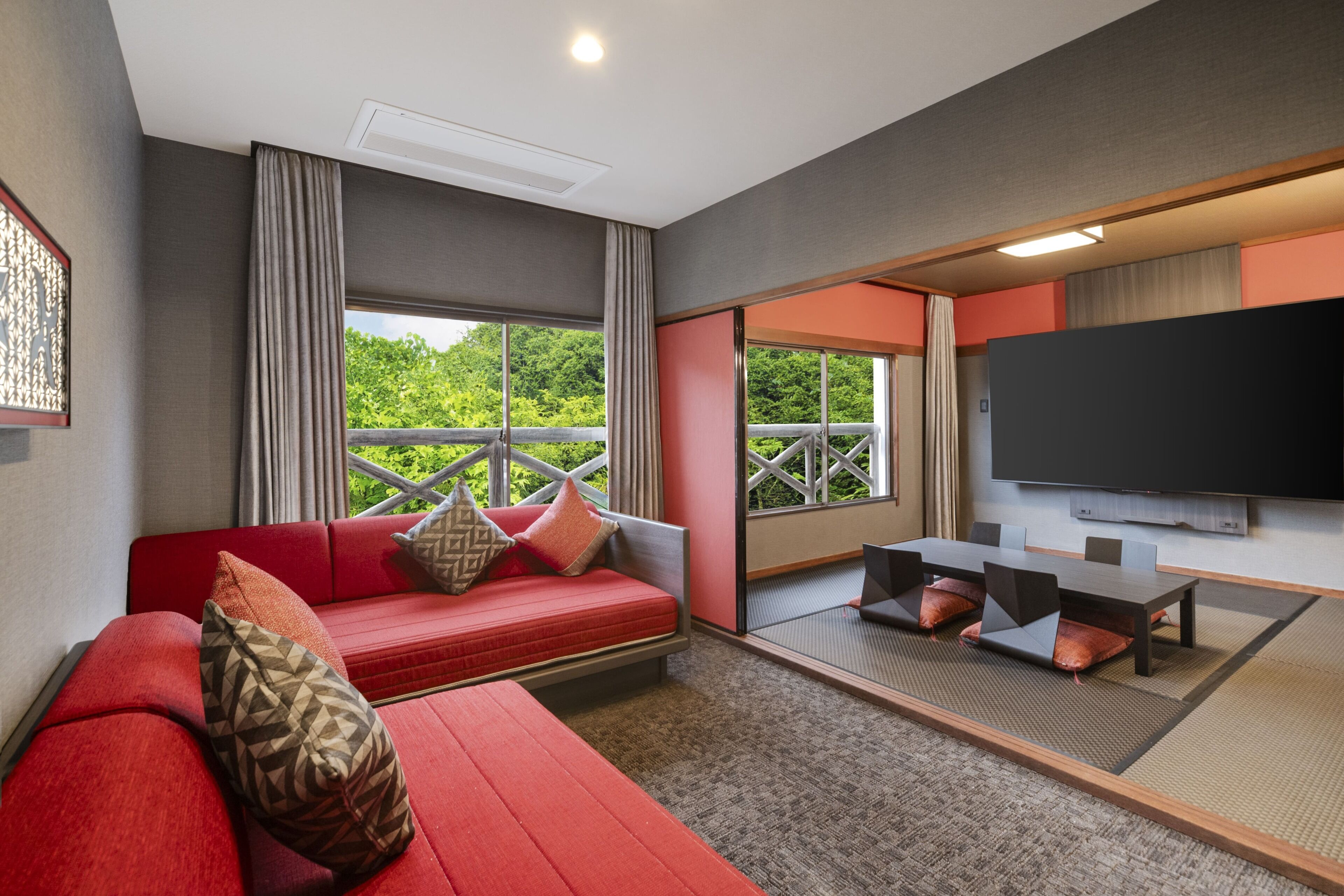 A modern room with a red sofa, a flat-screen TV, and a balcony with a view of greenery.