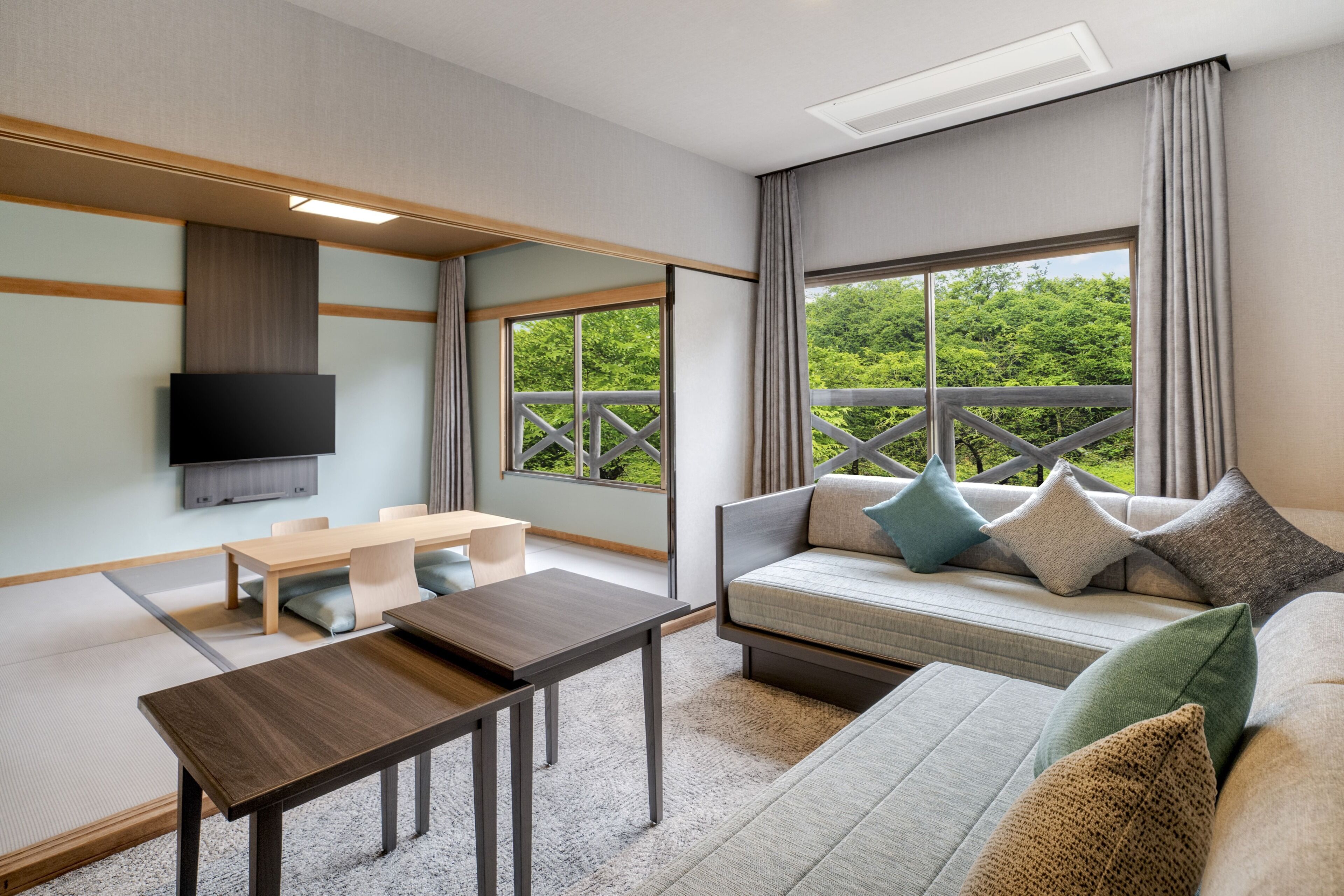 A modern room with a sofa, a TV mounted on the wall, a wooden table, and a view of greenery outside.