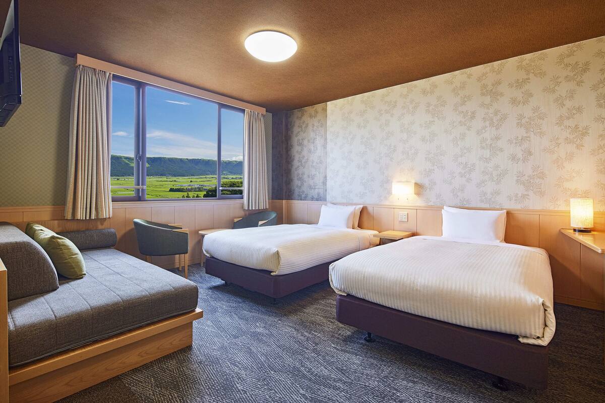 A hotel room with two beds, a sofa, a chair, and a large window with a view of the countryside.