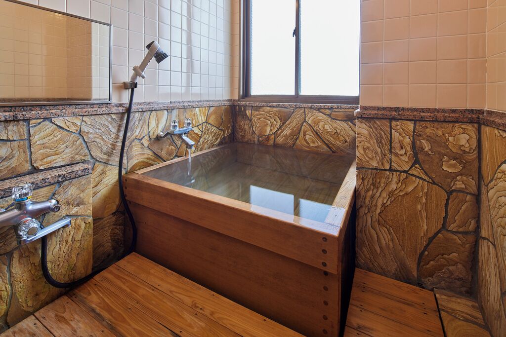 A stone-walled bathroom with a wooden bathtub, a window, and a shower head.