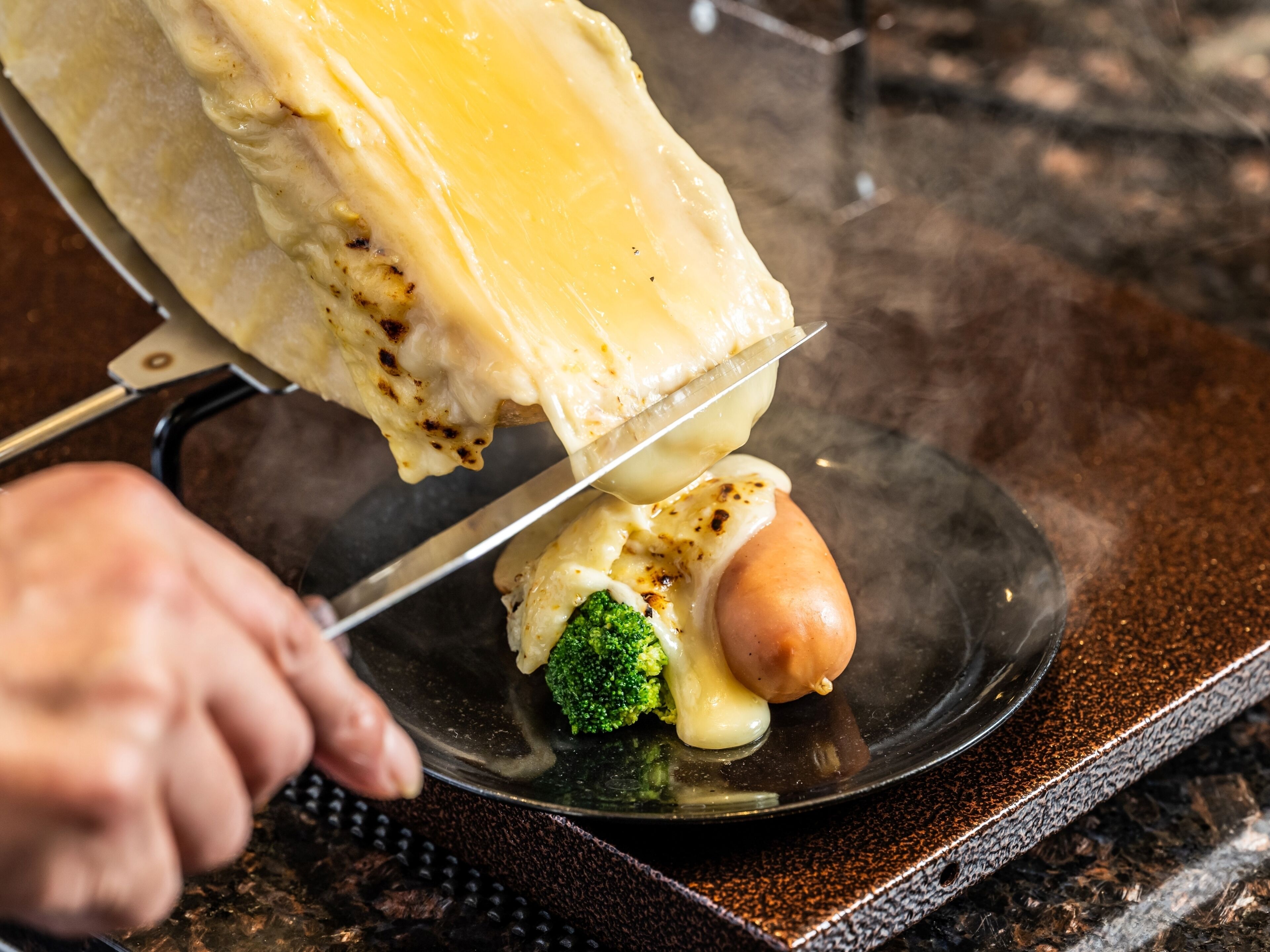 A person cutting a hot dog with melted cheese on a plate.