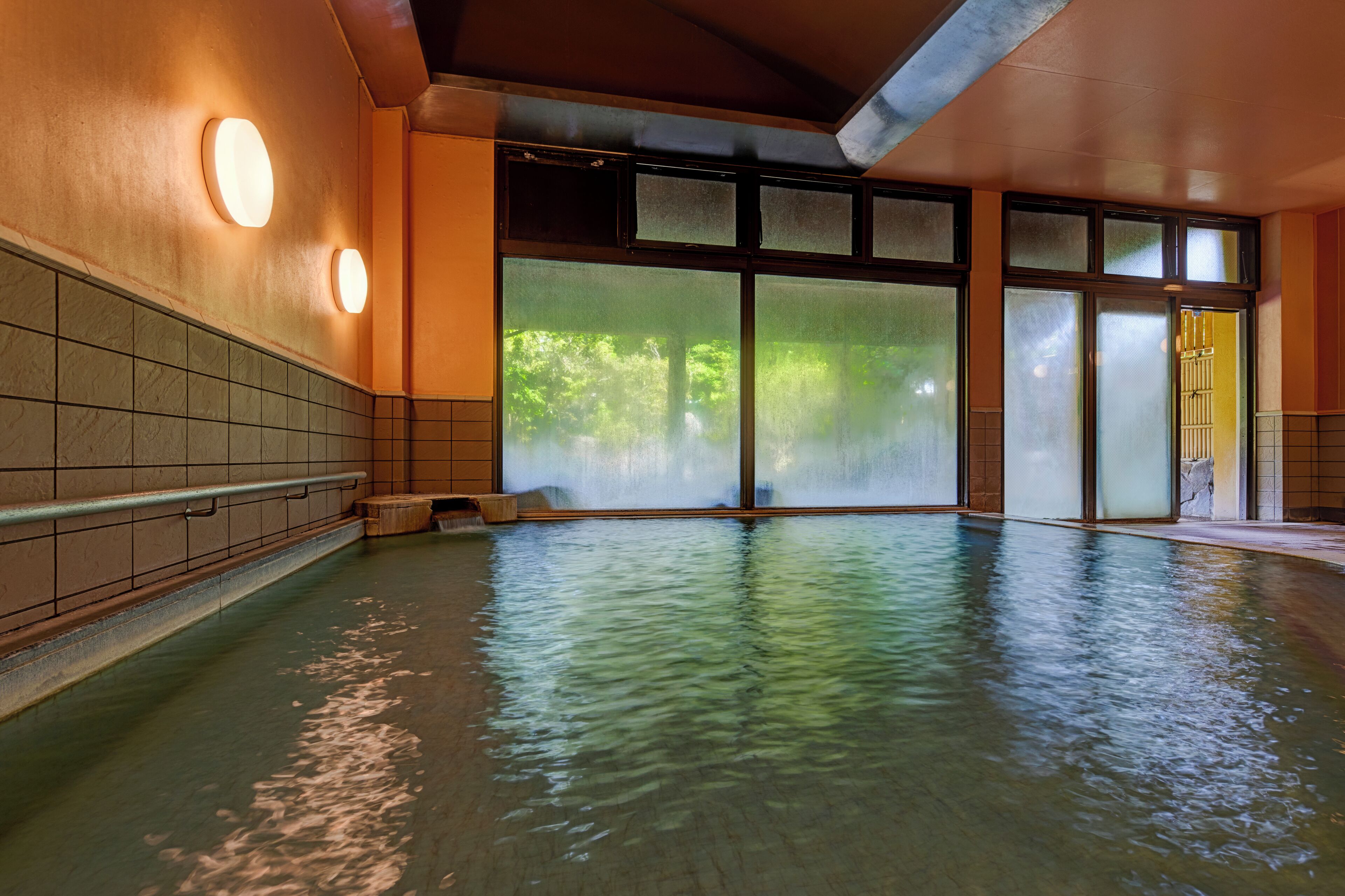 A large indoor swimming pool with clear glass walls, a tiled floor, and a wooden bench.