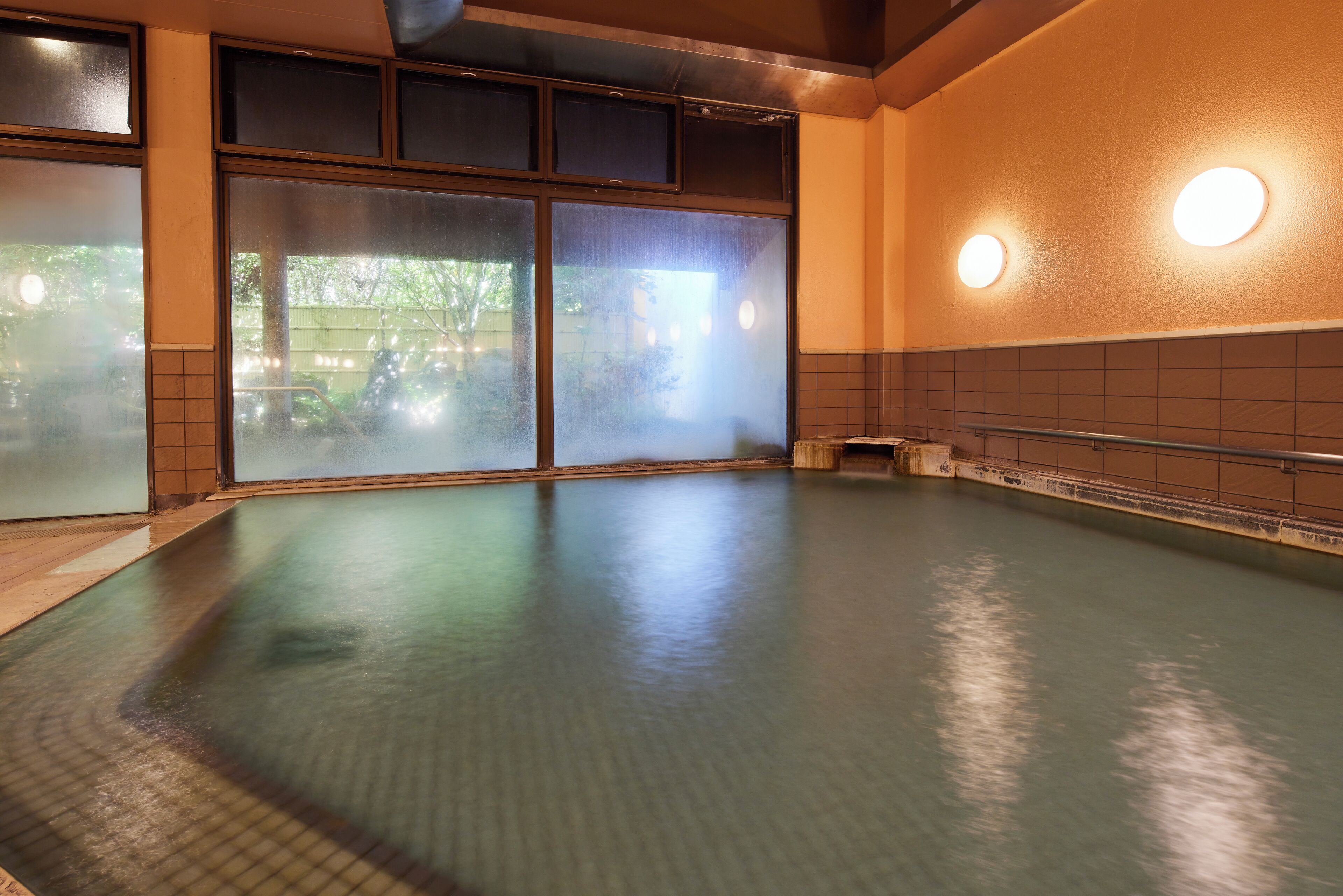 A large indoor swimming pool with clear glass walls, a tiled floor, and a metal railing along the edge.