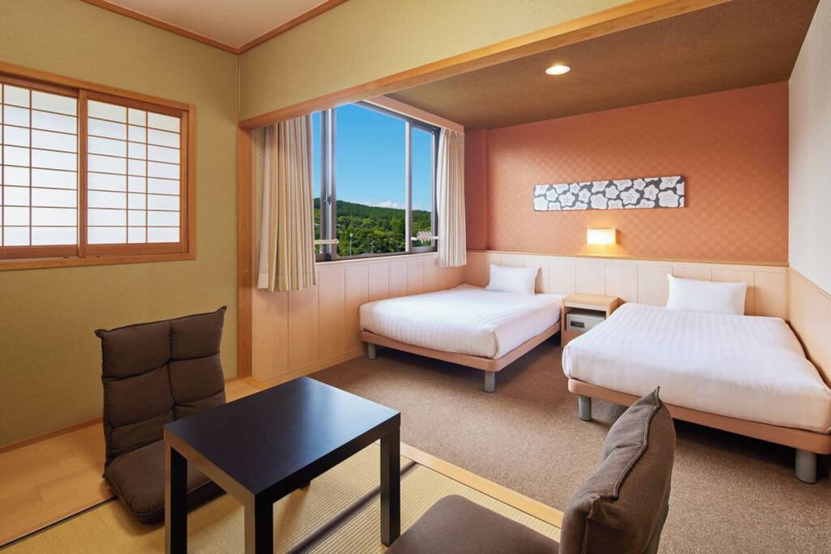 A hotel room with two beds, a small table, a chair, and a view of greenery through the window.