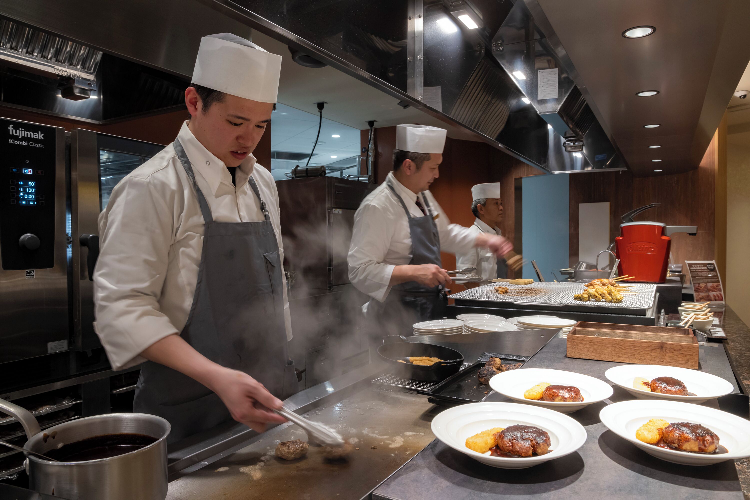Chefs preparing food in a professional kitchen with stainless steel appliances and a large cooking surface.