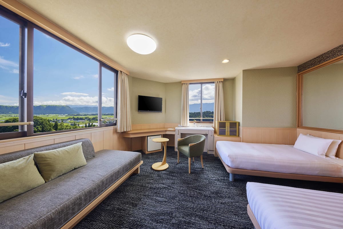 A hotel room with a sofa, two beds, a desk, a chair, a TV, and large windows with a view of mountains.