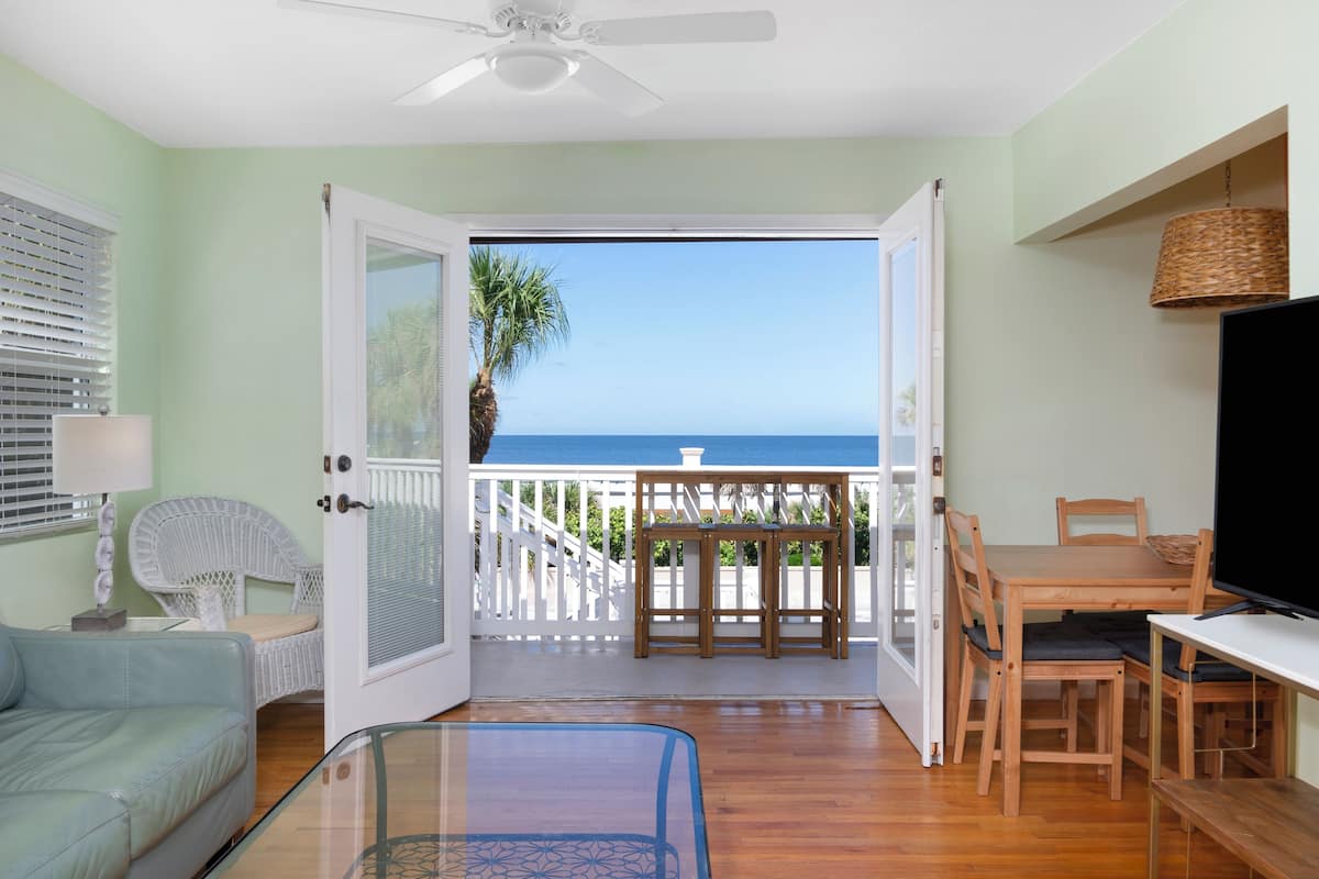 An open door leading to a balcony with a view of the ocean, a dining table, chairs, a television, and a wicker lamp.