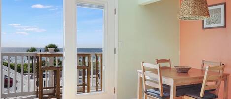 A dining area with a wooden table and chairs, a woven pendant light, and a view of the ocean through a sliding glass door.