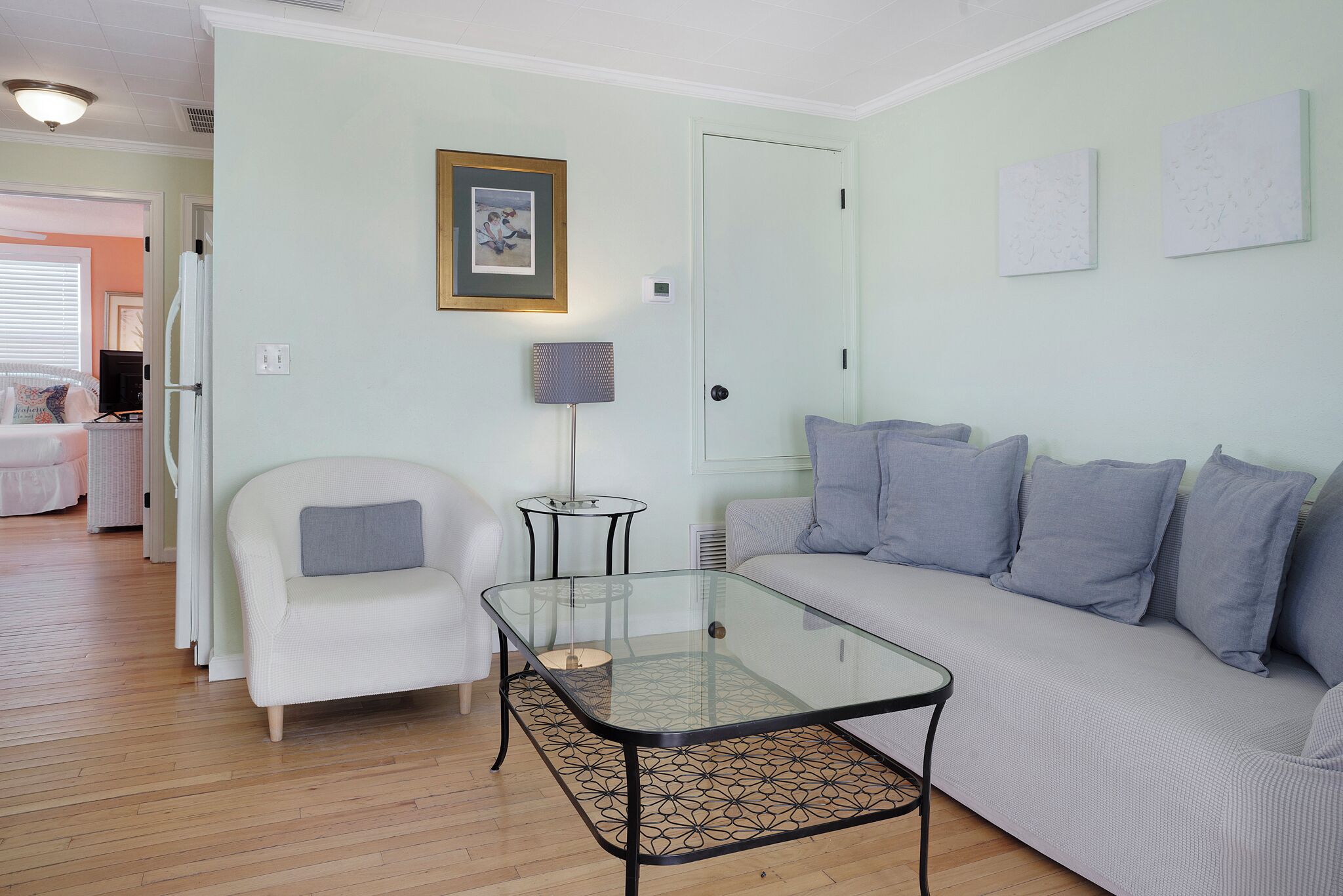 A living room with a glass-top coffee table, a white sofa with grey cushions, a white armchair, and a small side table with a lamp.