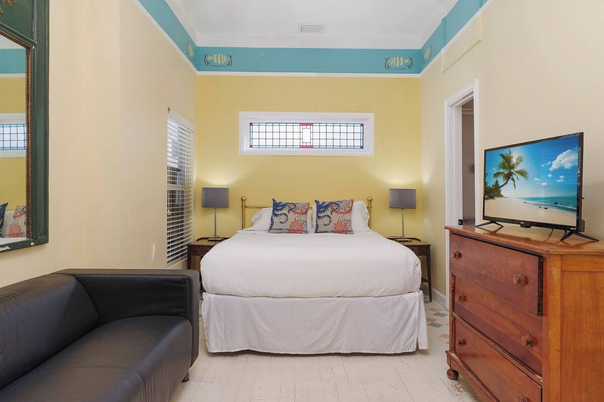 A bedroom with a bed, bedside lamps, a dresser, a TV, and a window with blinds.
