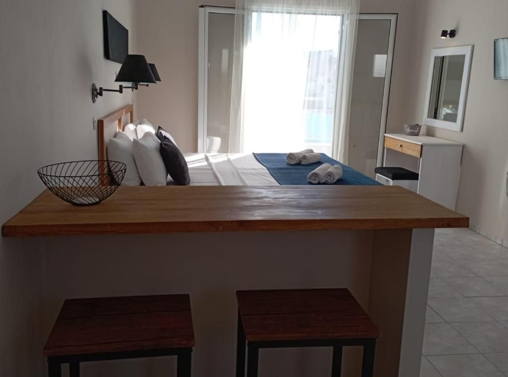 Quarto de hotel com secretária de madeira, dois taburetes, cama com roupa de cama branca, pequena mesa de cabeceira, luminária na parede, espelho e janela com cortinas.