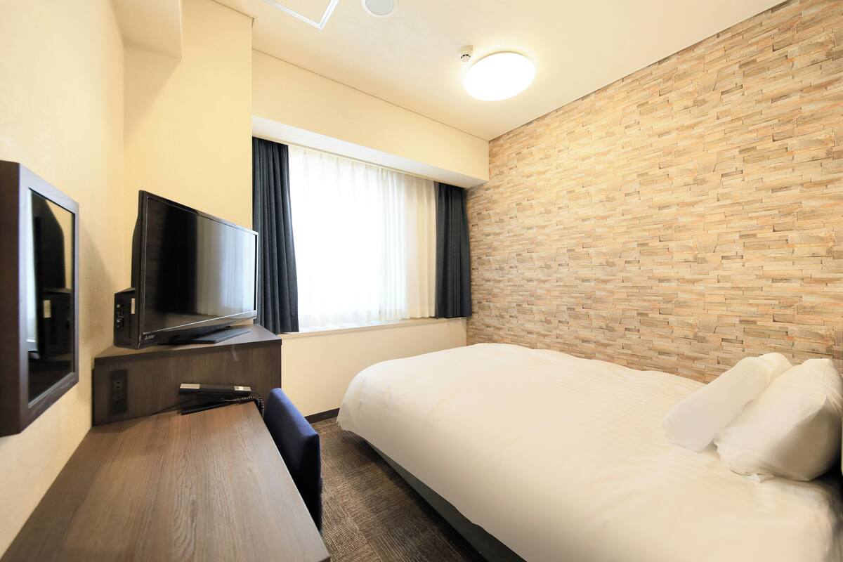 Una habitación de hotel con una cama, un escritorio con televisor de pantalla plana, una ventana con cortinas y una pared con diseño de ladrillos.