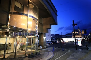 A hotel entrance with a glass facade, a visible reception area, and a street view with parked cars and a traffic light. - Hotel Nagano Avenue (Nagano)