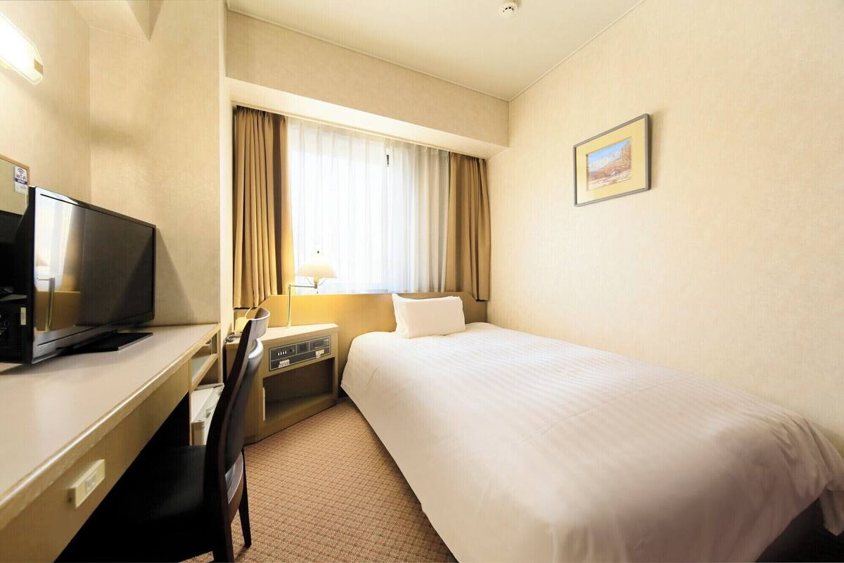 Una habitación de hotel con cama, escritorio con televisor, lámpara y ventana con cortinas.