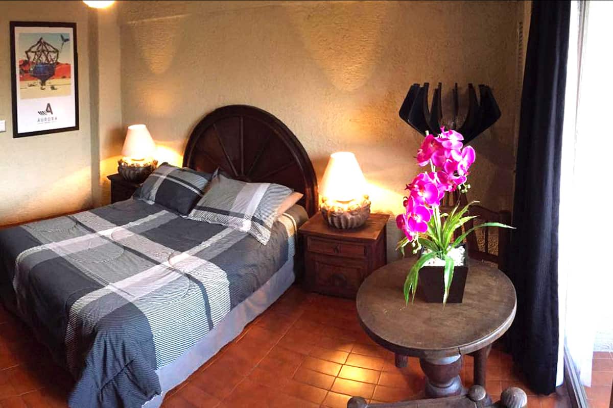 A hotel room with a bed, bedside lamps, a small table with a flower vase, and a television on the wall.