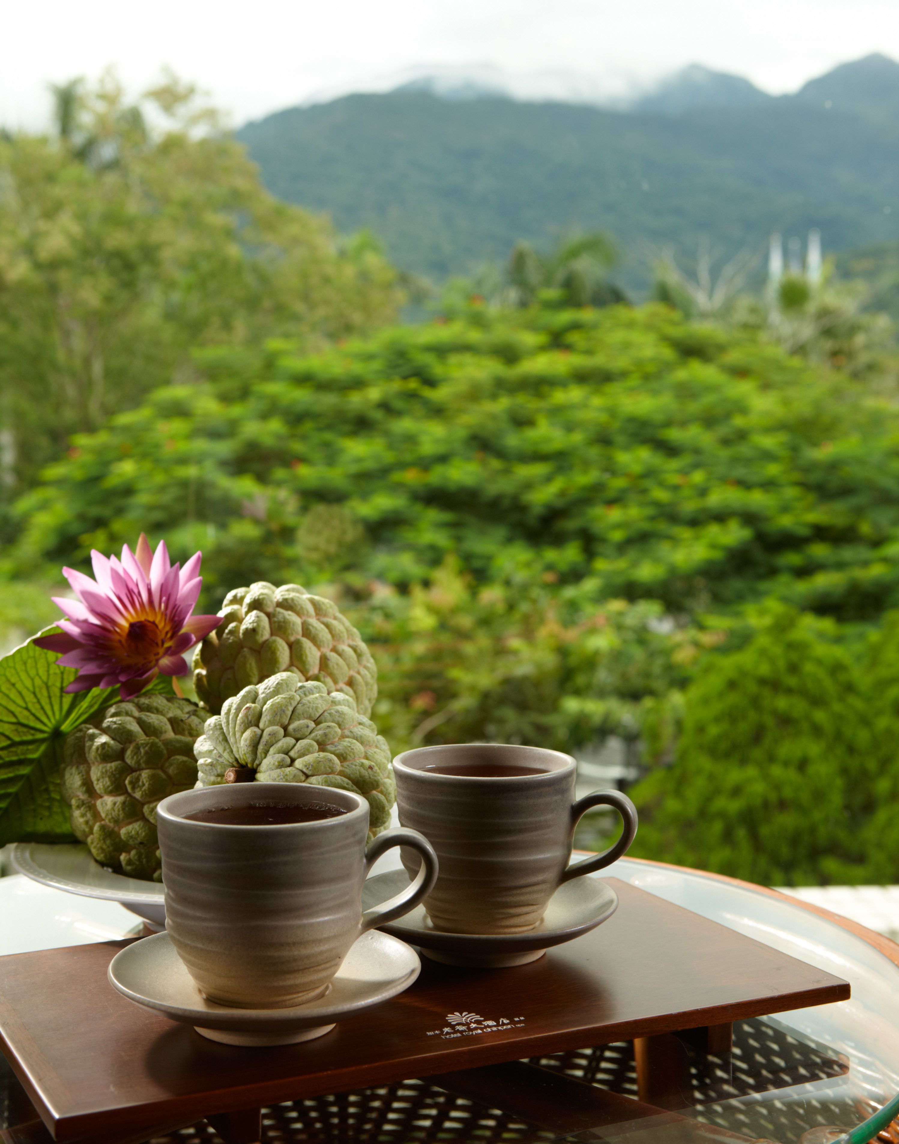 A table with two ceramic cups and a flower, set against a backdrop of lush greenery and mountains.